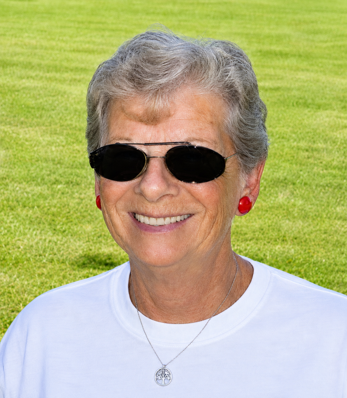 Portrait of Ann Grunewald, smiling and wearing sunglasses and a white shirt, outdoors on a green lawn